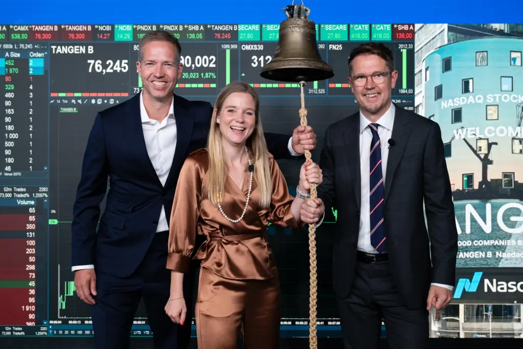 Nina Bergman, Fredrik Lofgren and Per Skånberg pose joyfully at the Nasdaq listing ceremony for Tången B. Nina is holding the exchange bell, with trading screens and the Nasdaq logo visible in the background.