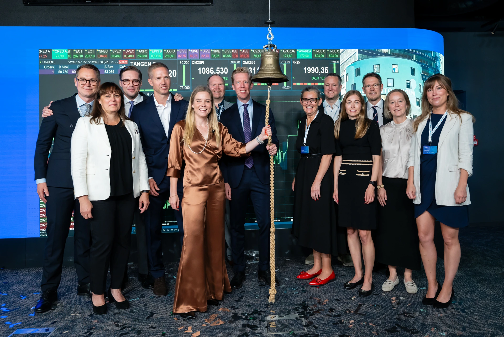 Nina Bergman and Per Andersson ring the stock exchange bell surrounded by employees and board members from Tången at the Nasdaq listing ceremony for Tången B. Trading screens displaying stock prices are visible in the background.