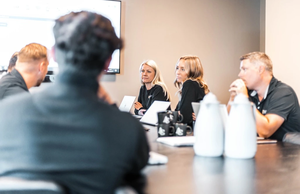 Five people sitting around a conference table, listening. A presentation screen is visible in the background.