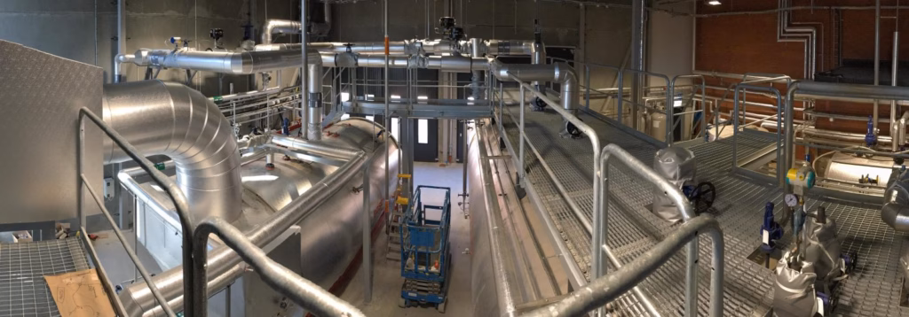 Interior of an industrial boiler plant showing large insulated boilers, metal pipes, valves and elevated walkways.